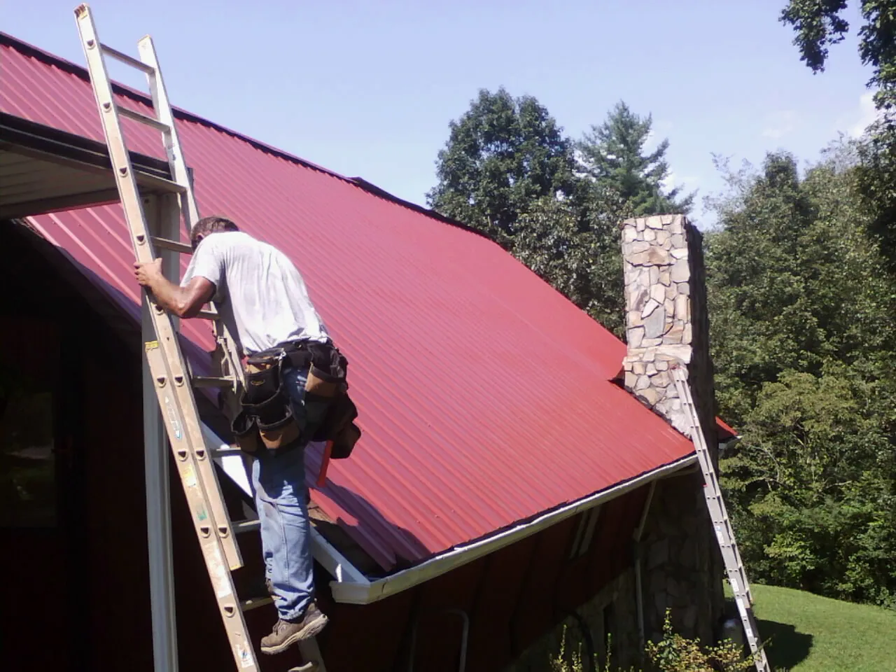 Roofer on a ladder beside a red metal roof and stone chimney — Soffit & Fascia Repair by Roofing Brothers in Steilacoom